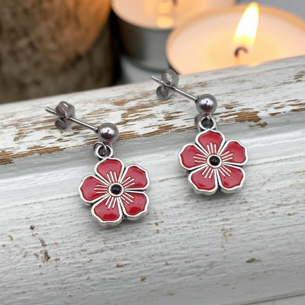 A pair of silver flower drop earrings with red painted petals and a small black centre, displayed on a weathered white wooden surface with blurred candles in the background.