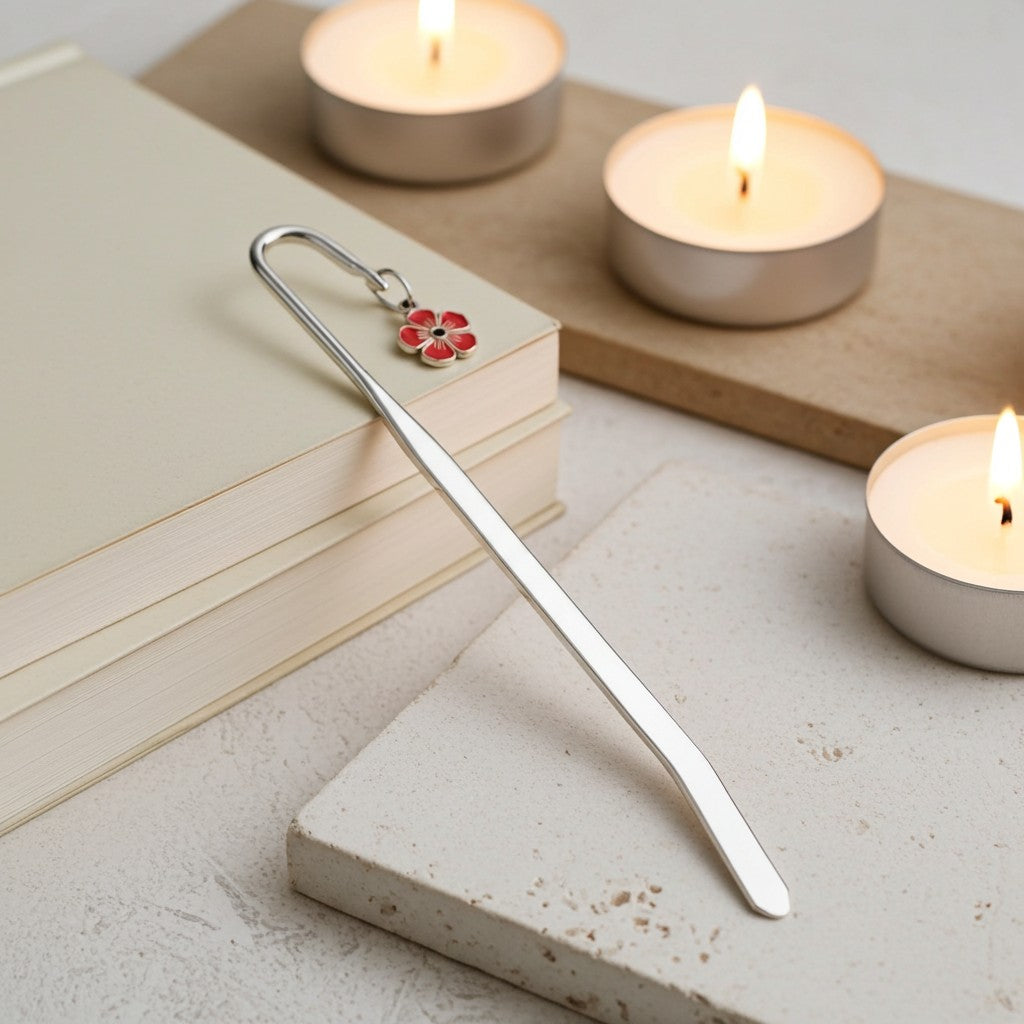 A silver metal bookmark with a small red and white flower charm rests on a stack of cream-coloured books, styled with lit tea light candles on a neutral stone surface.