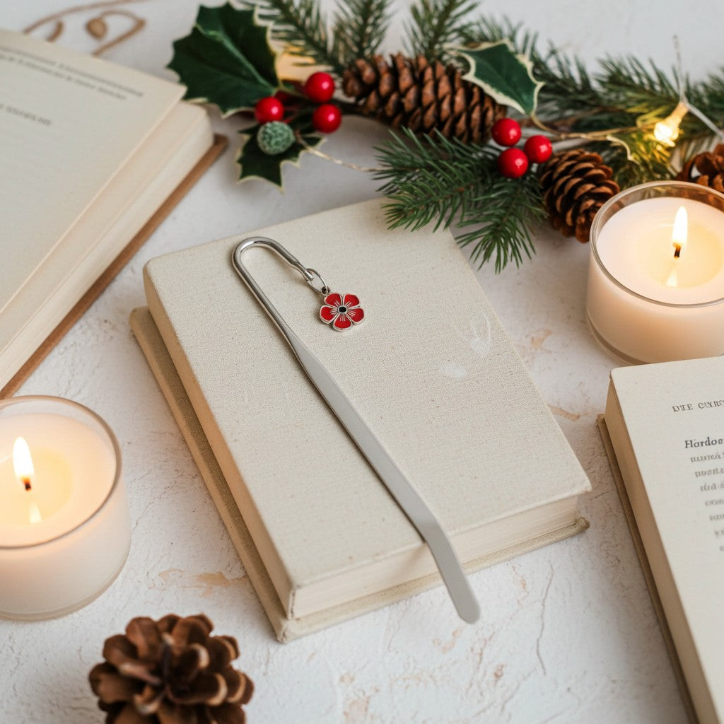 A silver metal bookmark with a small red and green floral charm rests on a closed cream-coloured book, styled with lit candles, pinecones, holly, and other books on a light textured surface.