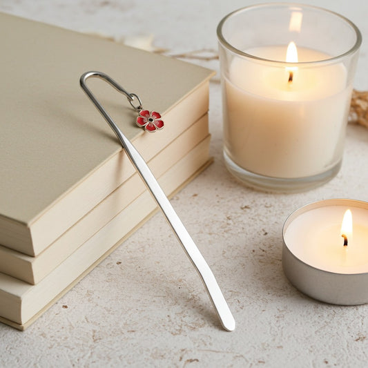 A silver metal bookmark with a small red and white flower charm rests on a stack of beige books next to two lit candles on a light stone surface.
