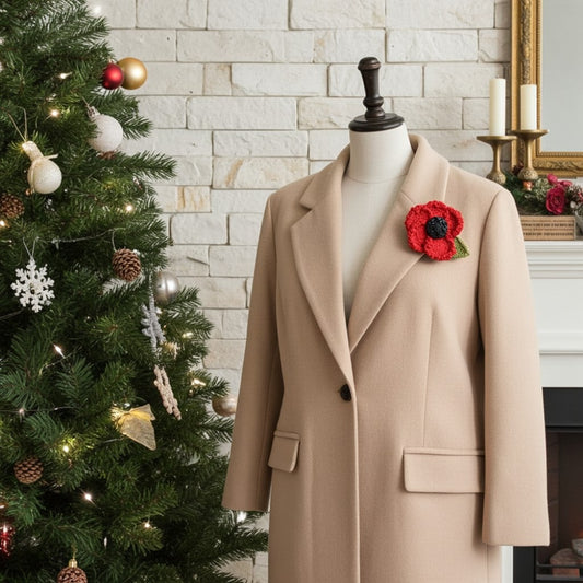 A beige wool coat displayed on a mannequin torso, styled with a red poppy brooch, set against a Christmas fireplace scene with a decorated Christmas tree and white brick wall.