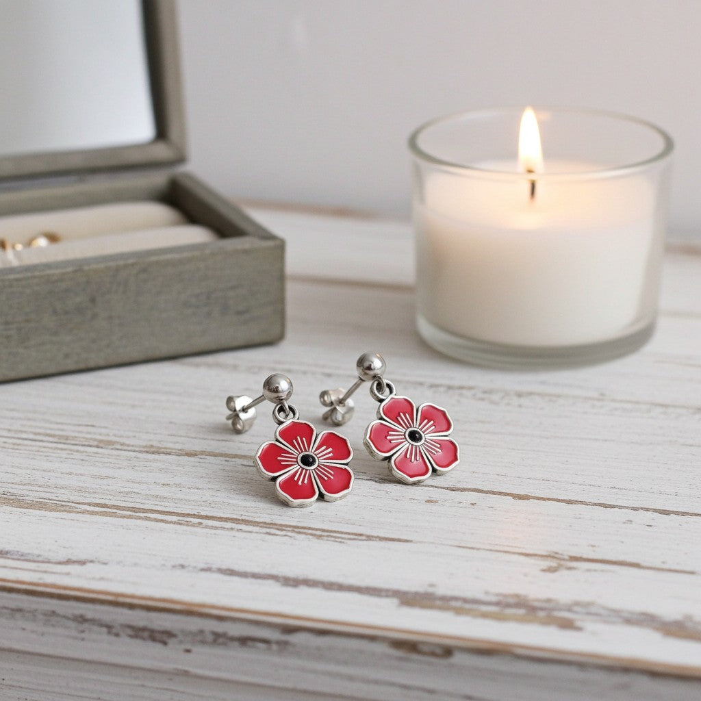 A pair of silver-toned flower-shaped earrings with red hand painted petals and black centres, displayed on a white wooden surface next to a lit white candle and an open jewellery box.