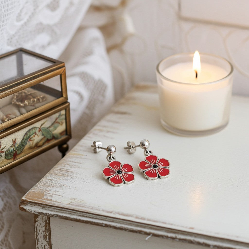 A pair of silver-tone flower dangle earrings with red painted petals and black centres, resting on a distressed white wooden table next to a lit white candle and a small decorative jewellery box.