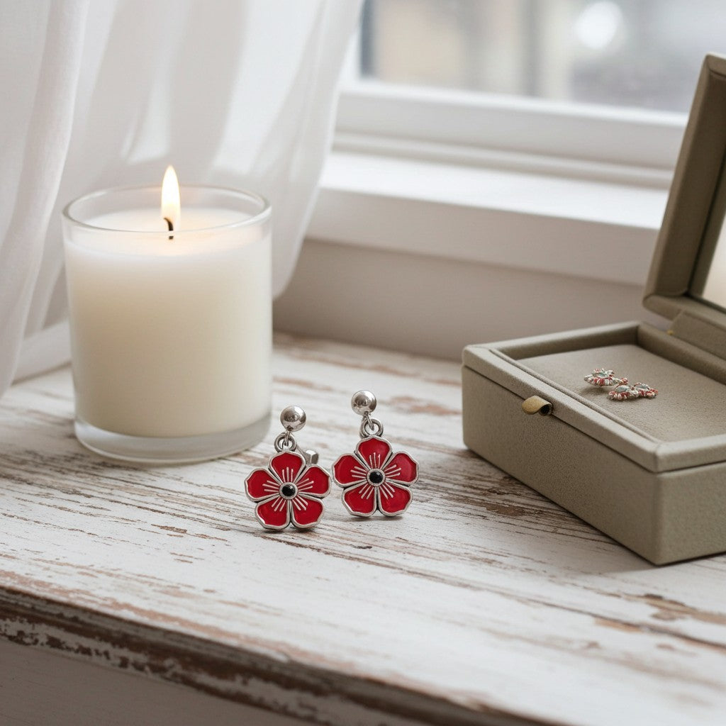 Red flower-shaped drop earrings with silver posts and black centers, displayed on a rustic white wooden table next to a lit candle and an open jewellery box.