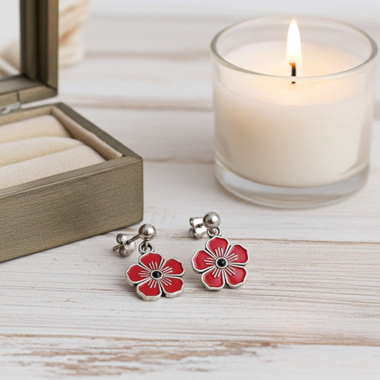 A pair of silver flower-shaped drop earrings with red painted petals and black centres, displayed on a light wood surface next to an open jewellery box and a lit white candle in a glass holder.