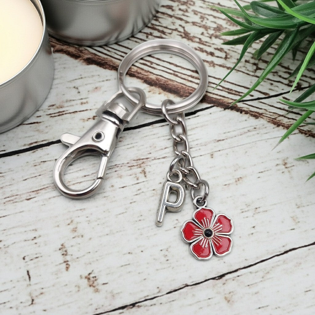 A silver-toned metal keychain with a lobster clasp, split ring, a hanging letter 'P' charm, and a red flower charm, displayed on a rustic white wooden surface with decorative props.