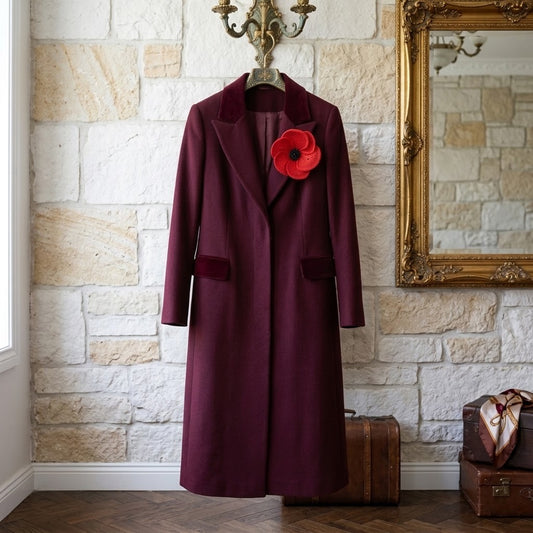 A long burgundy wool coat with velvet lapels and a red poppy flower pin, hanging on a wall hook in front of a stone wall.