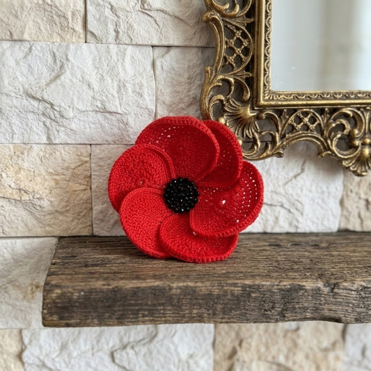 A red crocheted poppy flower brooch with a black centre, displayed on a rustic wooden shelf against a light stone wall with an ornate gold mirror frame.