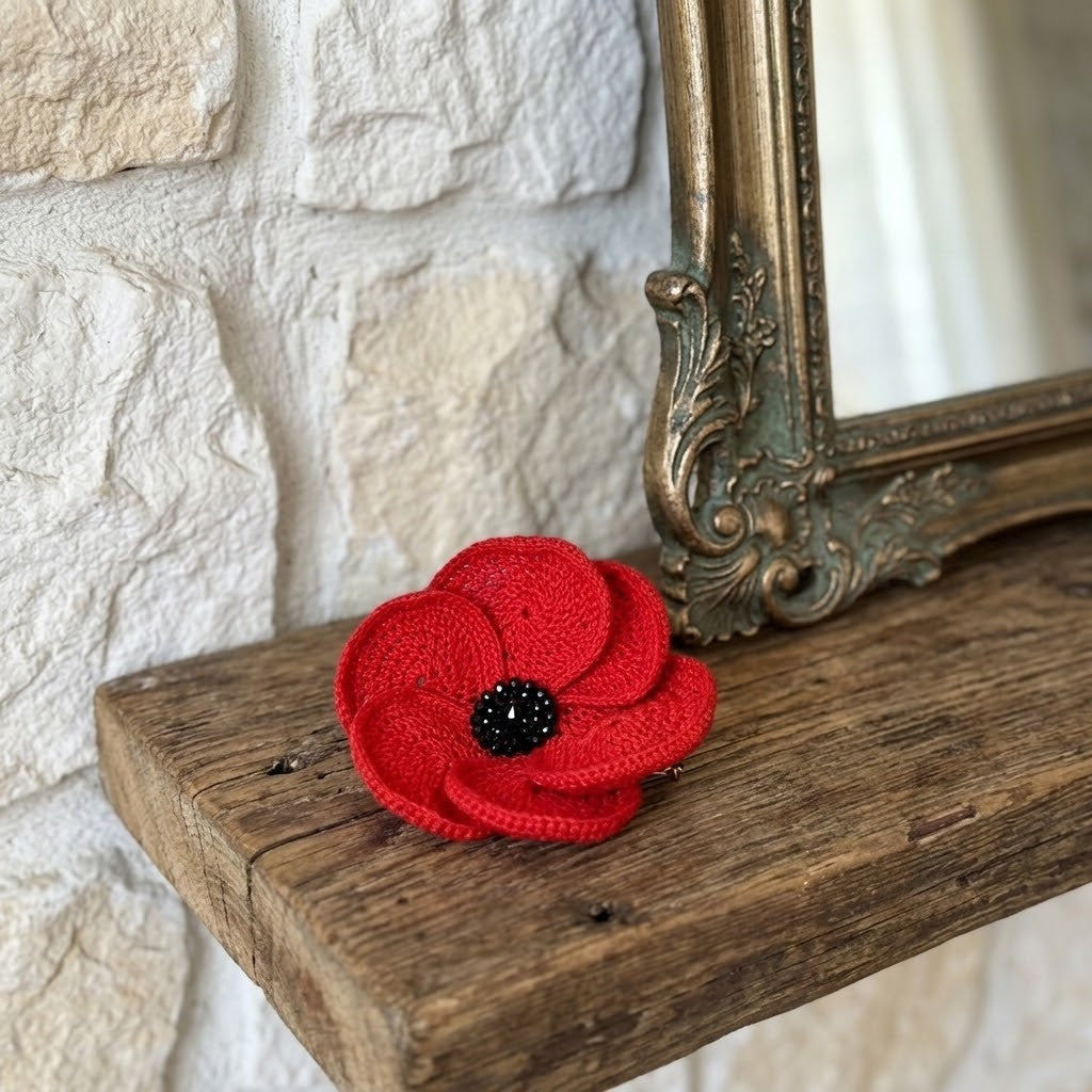 A bright red crocheted poppy flower brooch with a black centre sits on a rustic wooden shelf next to an ornate gold mirror against a white stone wall.