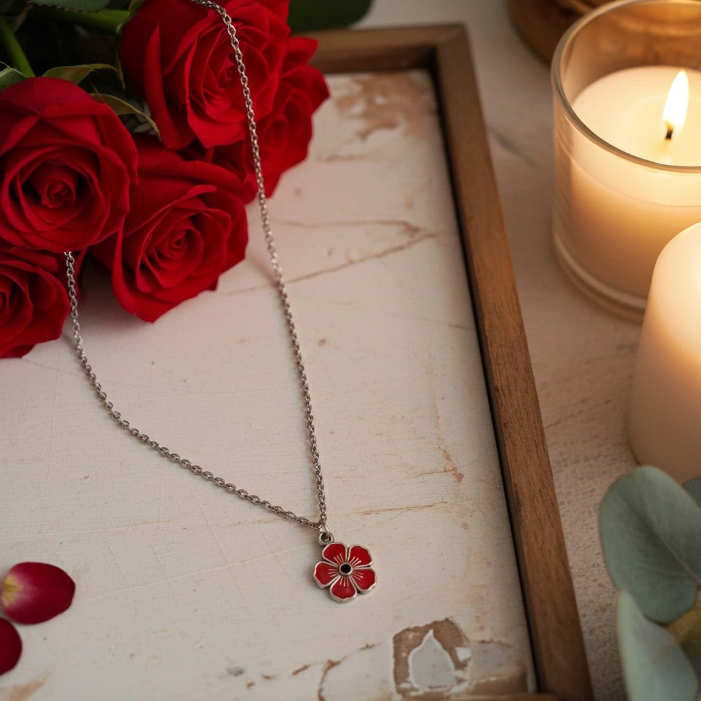A silver chain necklace with a red flower-shaped pendant, styled on a distressed white tray with red roses, candlelight, and a wooden frame.