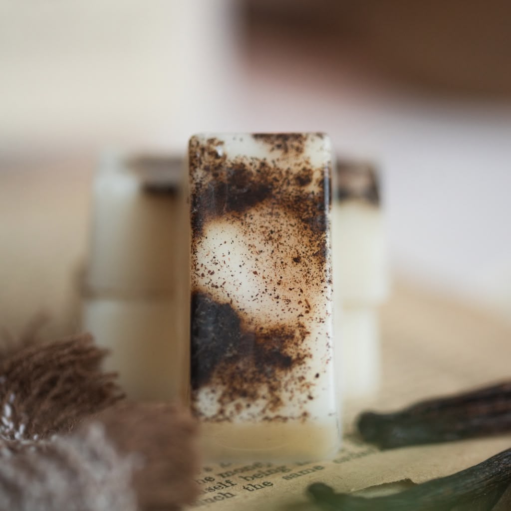 A rectangular bar wax melt with a marbled pattern of cream, white, and dark brown speckles, standing upright on a rustic surface with other soap bars blurred in the background.