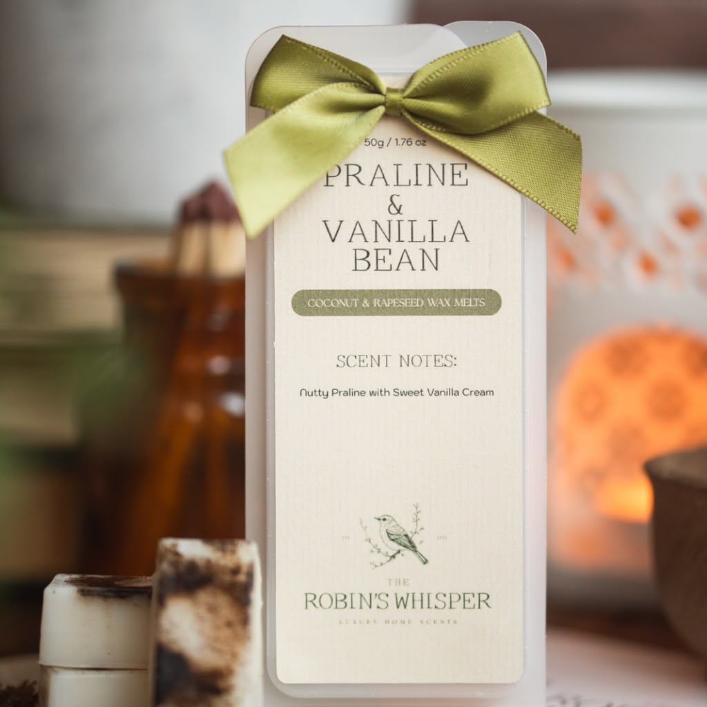 A wax melt package labeled 'Praline & Vanilla Bean' tied with an olive green satin bow, standing upright in a softly blurred warm-toned setting with candles and wooden decor in the background.