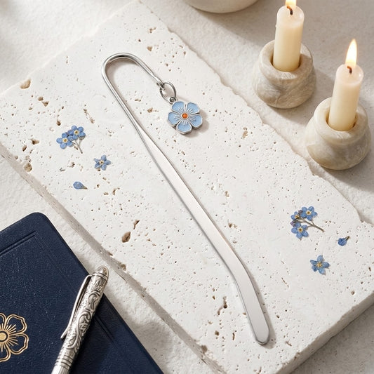 A silver metal bookmark shaped like a flower with blue enamel petals and a small gold centre, resting on a textured white stone slab with scattered blue flower petals, candles, and part of a dark notebook visible.