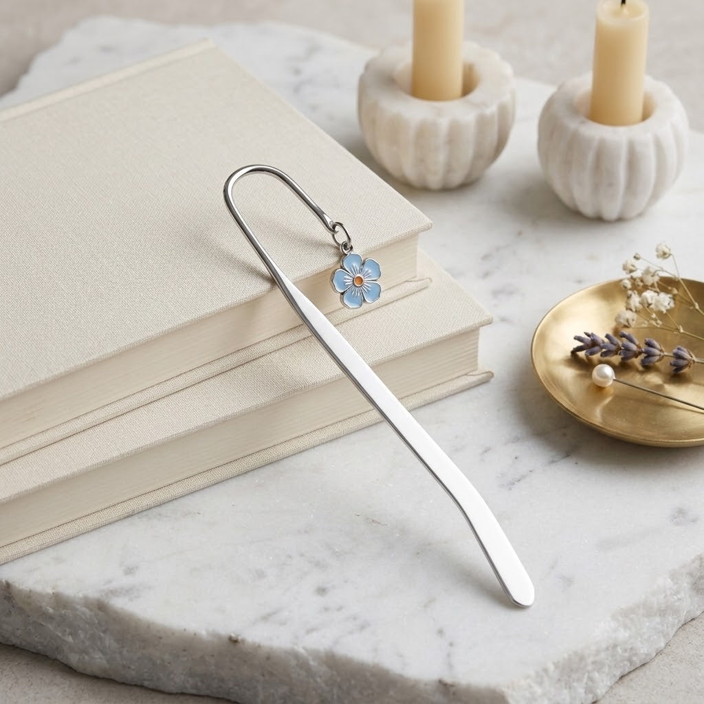 A silver metal bookmark with a small light blue flower charm rests on a stack of cream-coloured books on a white marble surface, with candles and a gold dish in the softly blurred background.