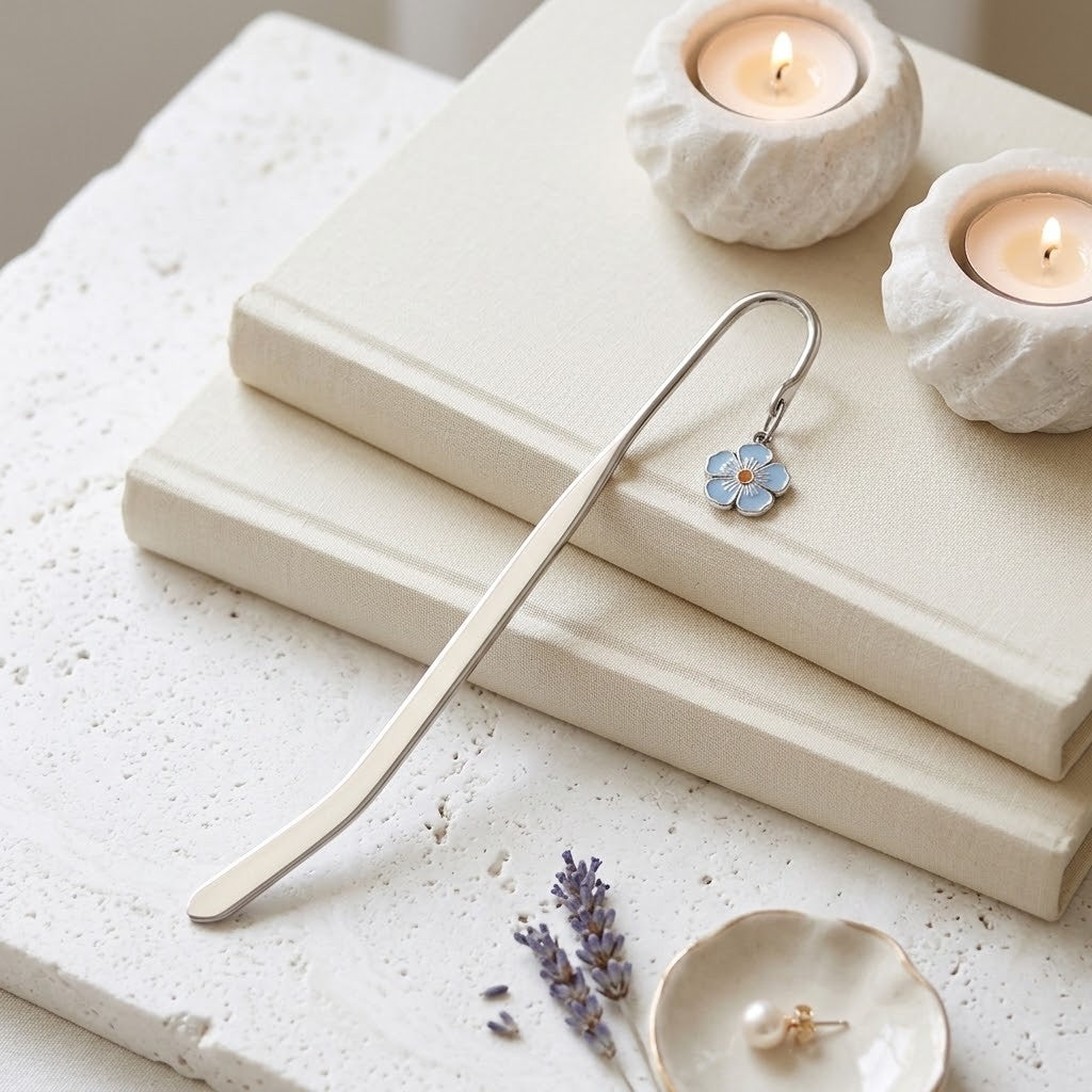 A silver metal bookmark shaped like a small blue flower, resting on stacked cream-coloured books with lit candles and dried lavender sprigs nearby.