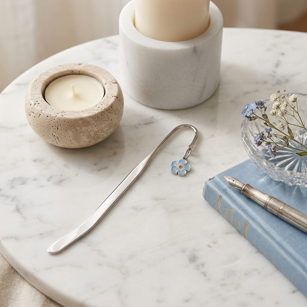 A silver metal bookmark shaped like a small blue flower with white petals, resting on a white marble tray alongside candles, a notebook, and a pen.