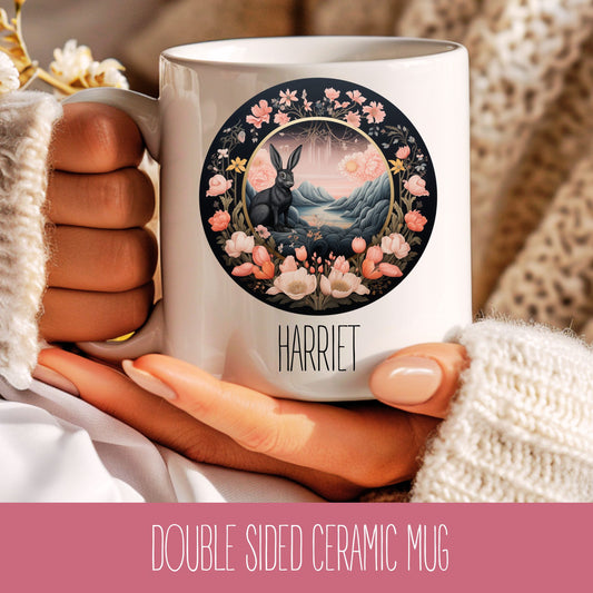 A ceramic coffee mug with a floral and animal design, featuring a rabbit, flowers, and a moon and stars motif, held in a person's hand.