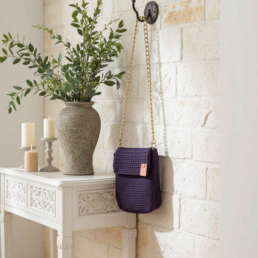 Purple woven bag with a gold chain hanging on a white brick wall next to a vase with greenery and candles.