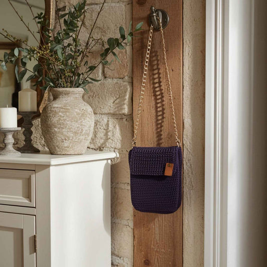 Purple woven bag with a gold chain hanging on a wooden door next to a stone wall and decorative items.