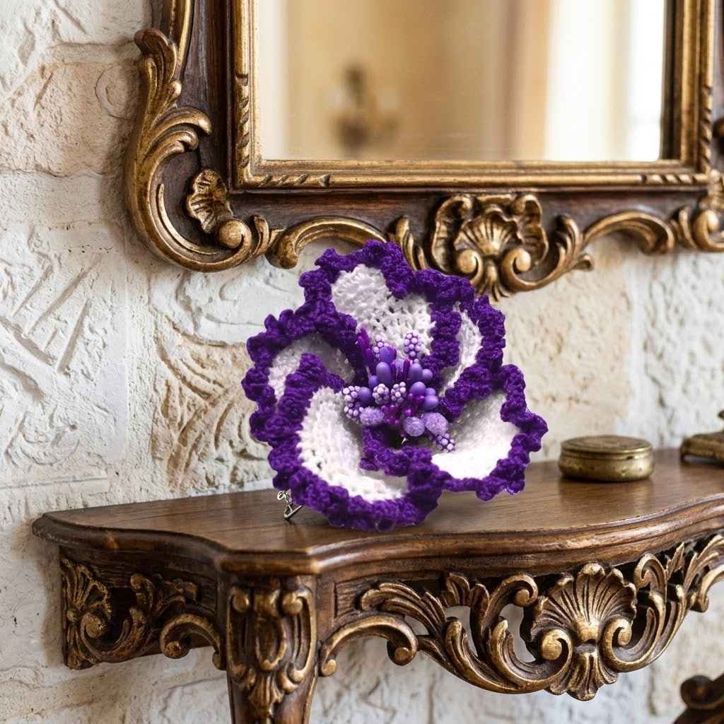 A crocheted flower-shaped brooch in purple and white, displayed on an ornate wooden console table beneath a matching decorative mirror.