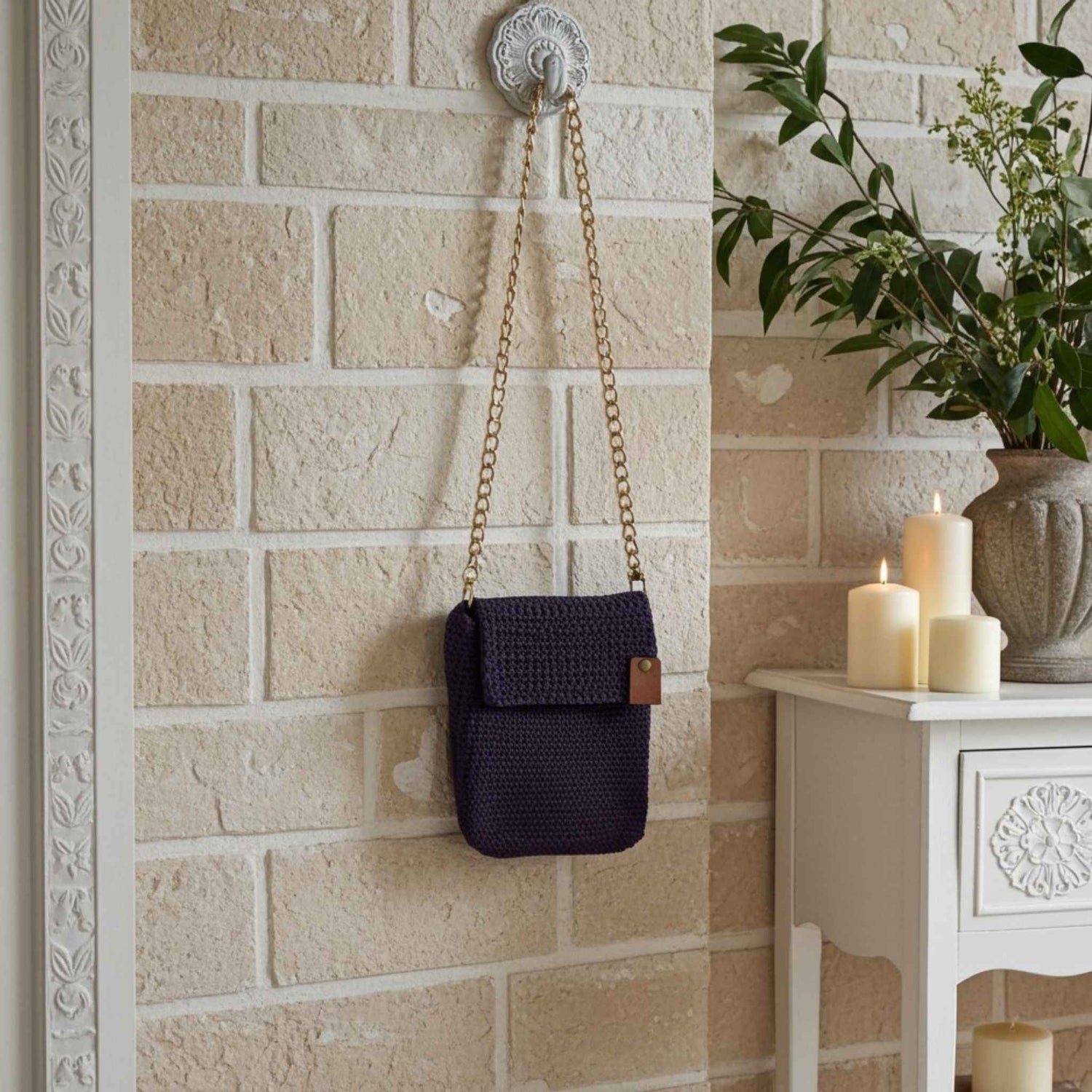 Purple woven bag hanging on a wall hook against a beige brick wall with a decorative white cabinet and plant in the background.