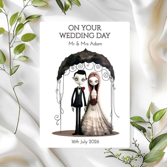 Wedding card with cartoon gothic couple under an umbrella, surrounded by green leaves on a white fabric background.