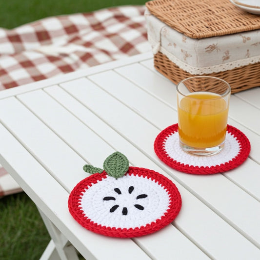 Two crocheted apple-shaped coasters on a white slatted table outdoors, with one coaster holding a glass of orange juice. A picnic blanket and wicker basket are blurred in the background.
