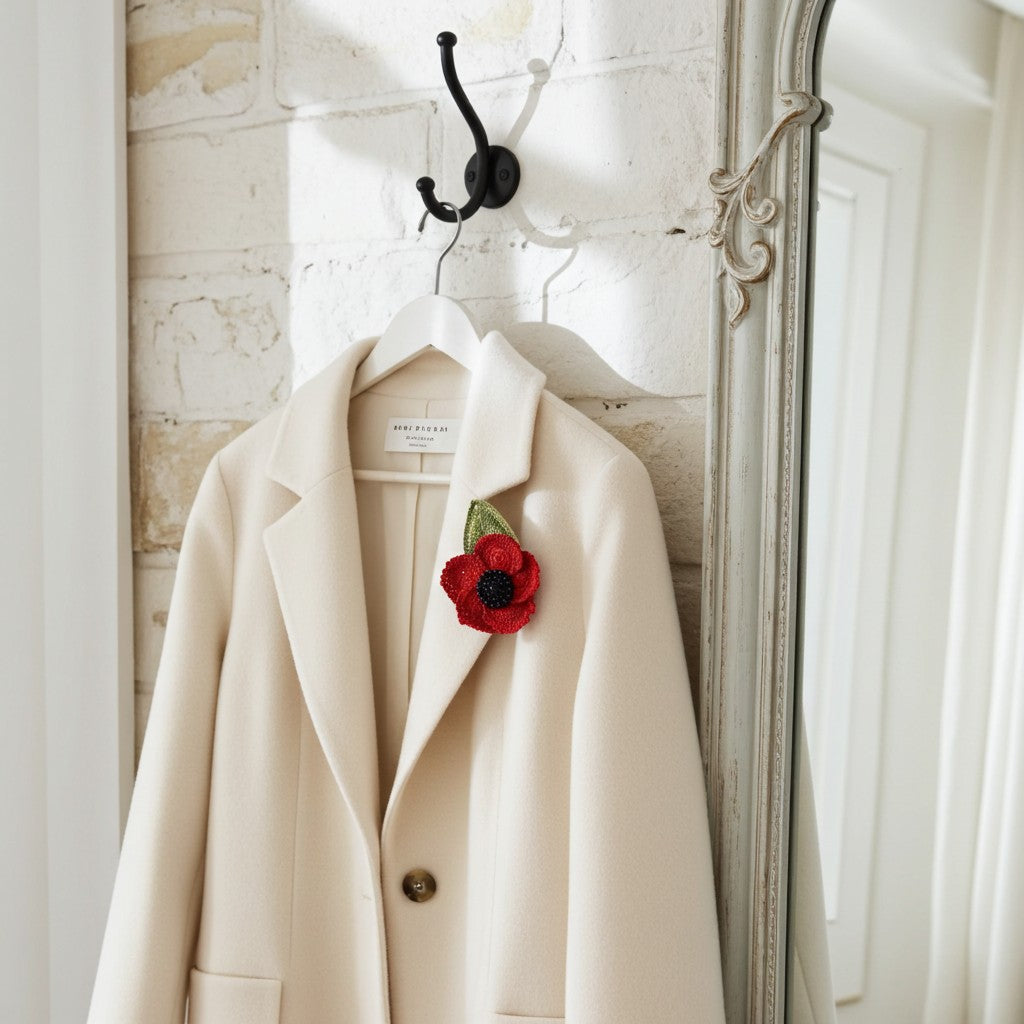 Beige coat with a red poppy pin on a hanger against a white wall.
