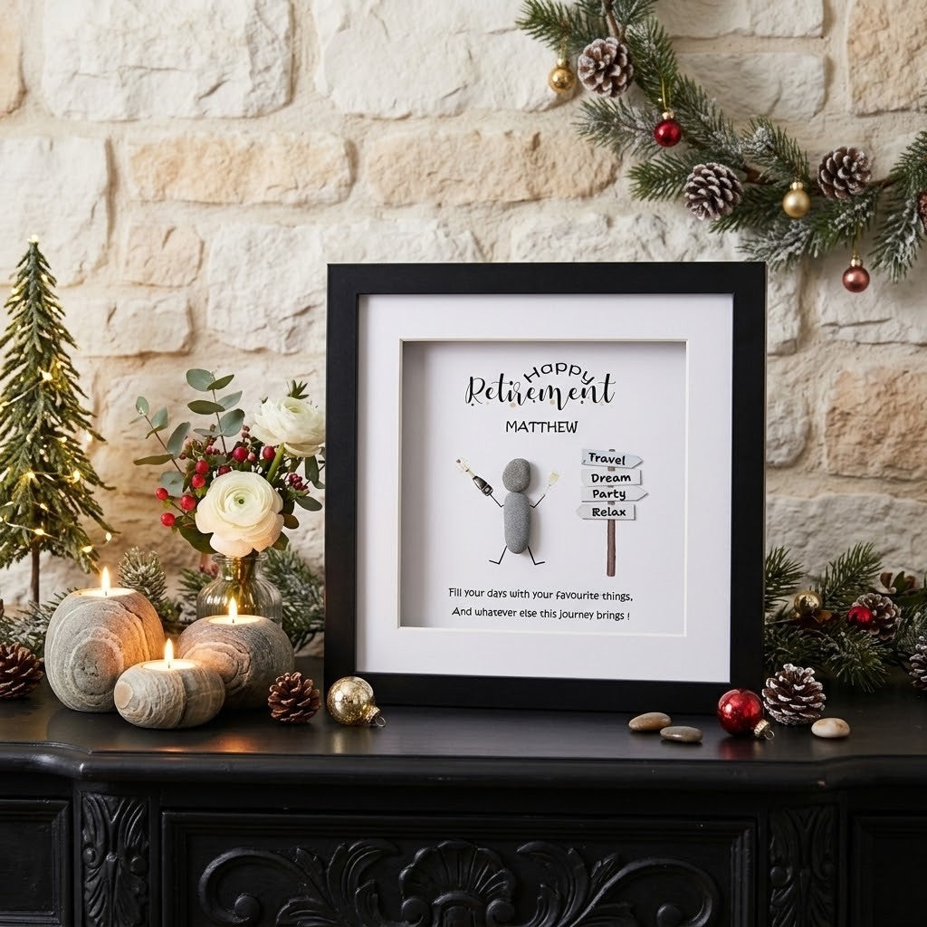 A framed retirement gift displayed on a black wooden chest against a light stone wall, decorated with pine garlands, candles, flowers, and ornaments. The black-framed print features a cartoon figure and the text 'Happy Retirement Matthew' with a sign reading 'Travel Dream Relax'.