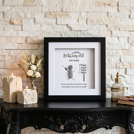 A framed personalised retirement gift displayed on a black carved console table against a light stone brick wall. The black frame contains a white background with a small stick-figure style character and the text 'HAPPY Retirement MATTHEW!' 