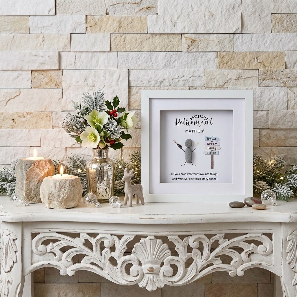 A framed retirement pebble art with a pebble art figure and personalised text displayed on a white console table against a beige stone wall, styled with candles, a floral arrangement, and small decorative items.