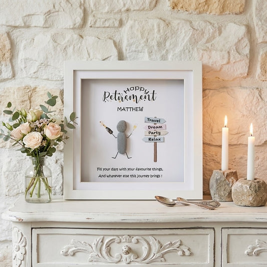 A framed personalised retirement gift displayed on a white wooden cabinet against a light stone wall. The white frame contains a printed message reading 'Happy Retirement Matthew' with a small stick-figure figure and signpost details, flanked by a vase of pink flowers and two lit candles.