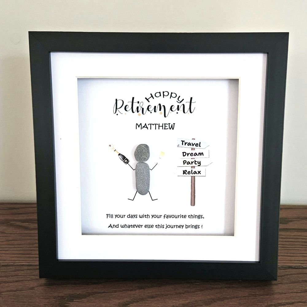 A framed retirement pebble gift with a black frame and white mat, featuring a personalised message for Matthew, a simple stick figure drawing, and a signpost with directions. The frame is displayed on a wooden surface against a light beige wall.