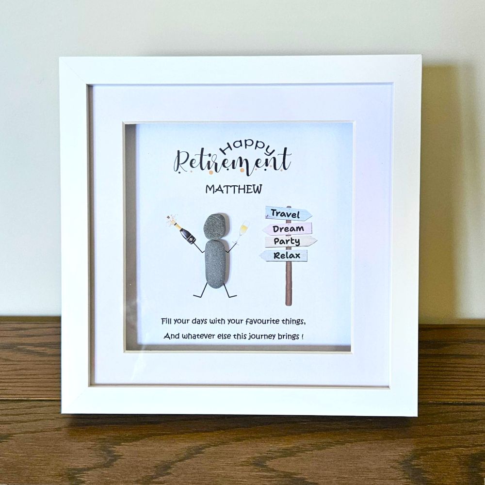 A white square framed pebble art gift leaning on a wooden surface against a light wall. The print says 'Happy Retirement Matthew' with a small stick-figure style figure and a signpost reading Travel, Dream Party, Relax, plus a short message below.