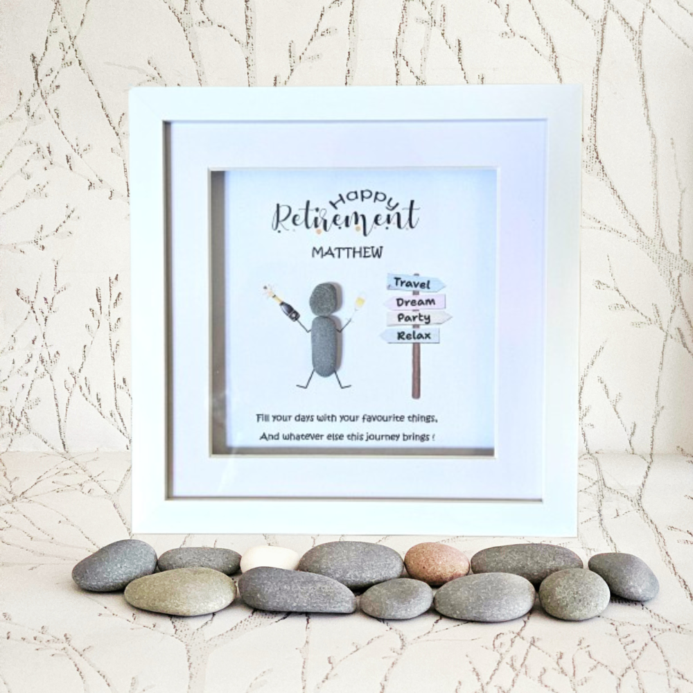 A framed personalised retirement gift displayed on a light surface with decorative stones. The white frame contains a printed card with the text 'Happy Retirement Matthew' and a simple illustration of a grey figure beside signposts reading 'Travel Dream Party Relax'.