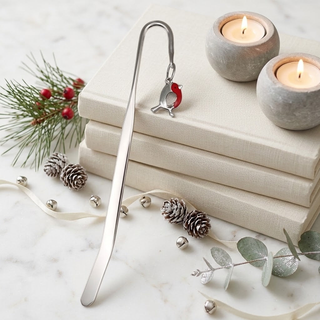 A silver metal bookmark shaped like a small robin bird with a red accent, displayed on a stack of cream-colored books with holiday decorations including pine branches, pinecones, silver bells, and lit candles.