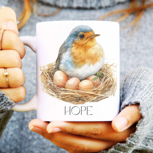 Mug with a robin and nest design labelled 'HOPE' held by hands.