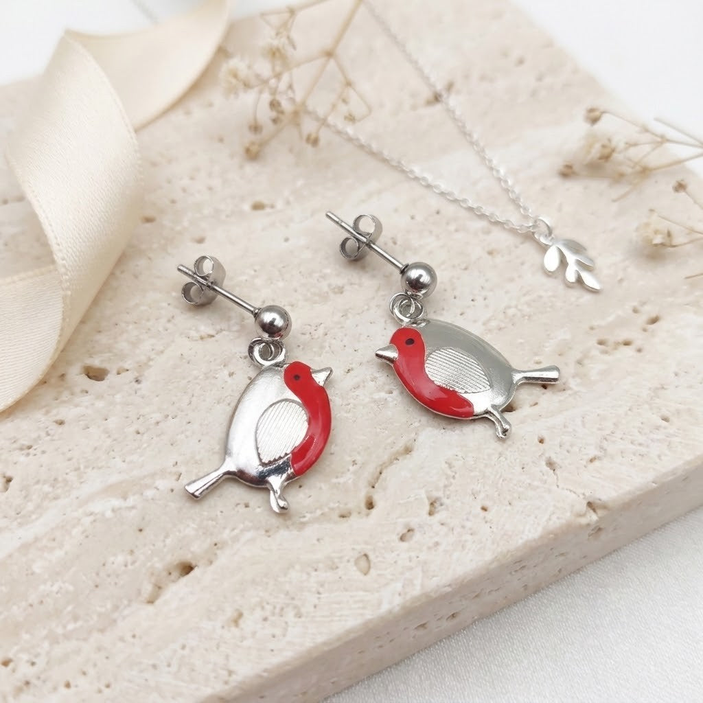 Silver robin stud earrings and a matching necklace displayed on a light stone surface with a cream ribbon and dried flowers. The birds are silver with bright red enamel accents.