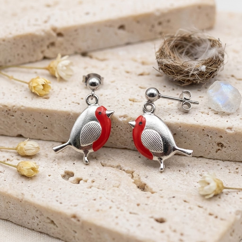 A pair of silver bird stud earrings with bright red accents, displayed on a textured beige stone slab with dried flowers and a small nest prop.
