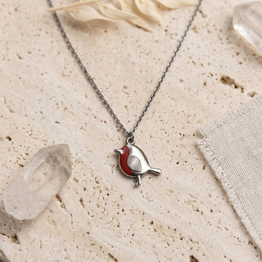 A silver bird pendant necklace with a red chest detail, displayed on a textured beige stone surface with decorative crystals and fabric.