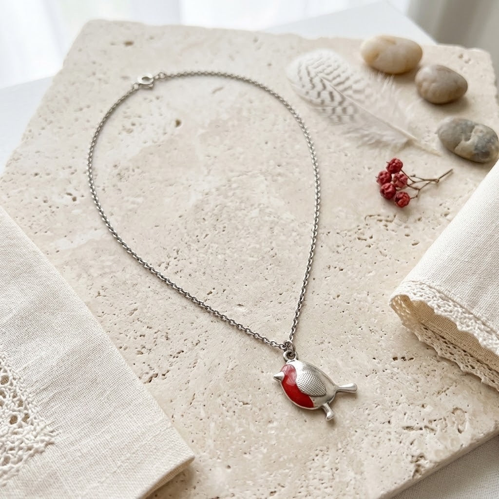 A silver chain necklace with a small bird pendant featuring a red chest, displayed on a textured beige stone slab with linen fabric, small stones, and dried red berries.