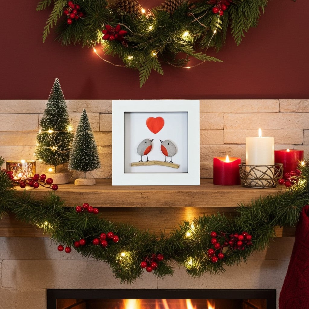 A square white-framed art print featuring two small robins facing each other beneath a red heart, displayed on a fireplace mantel surrounded by Christmas decorations including green garlands, red berries, candles, and miniature trees.