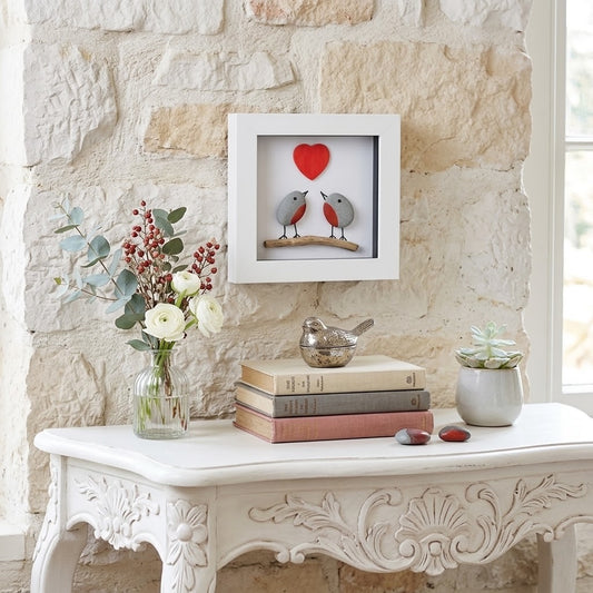 A white square shadow box wall art piece featuring two small birds facing each other beneath a red heart, displayed on a white carved console table against a rustic stone wall.
