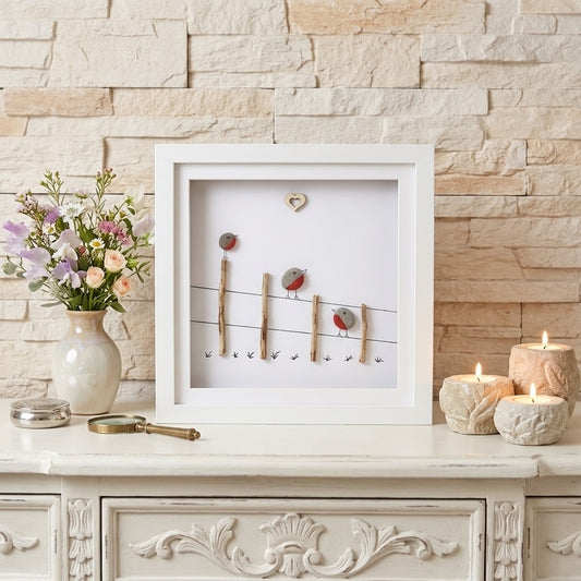 A framed decorative wall art piece displayed on a white cabinet against a beige stone wall. The artwork features small birds perched on wire-like lines with tiny hooks, set in a white frame.