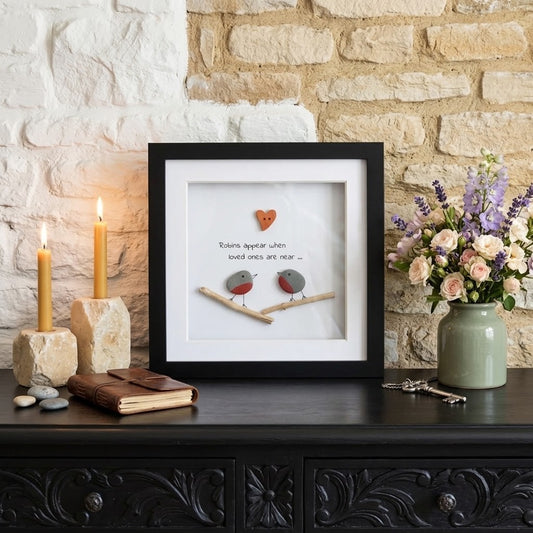 A framed shadow-box art print sits on a dark carved wooden console table against a white and beige stone wall. The print features a small orange heart, two tiny birds on branches, and the text 'Romance begins when loved ones are near.