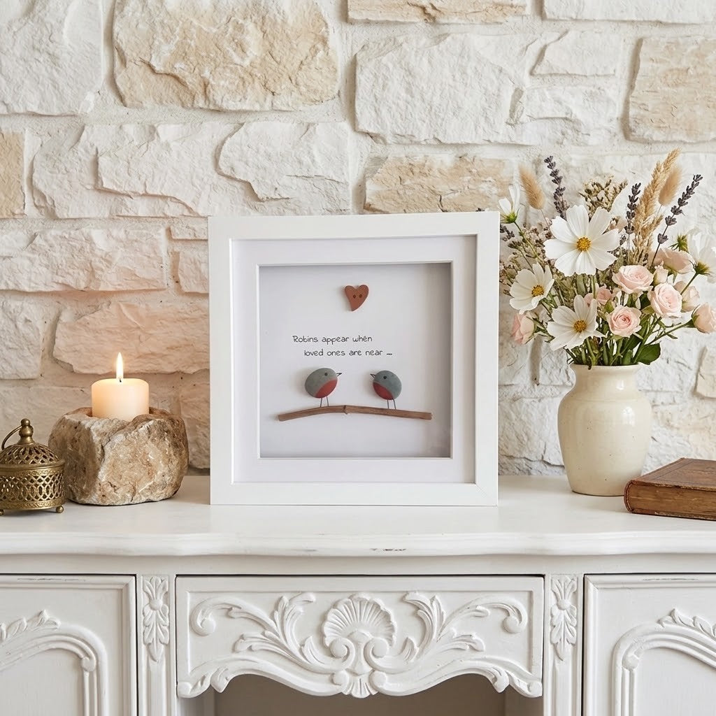 A white square shadow box frame displayed on a white console table against a light stone wall. The frame contains a small heart, a branch with two small round stones, and handwritten text.