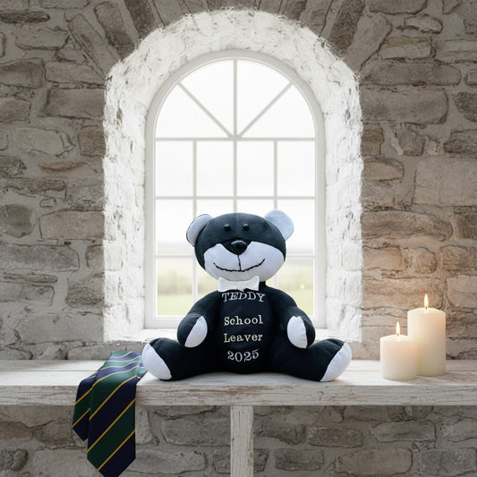 Black and white teddy bear with a school tie and 'School Leaver 2025' text on a stone wall background.