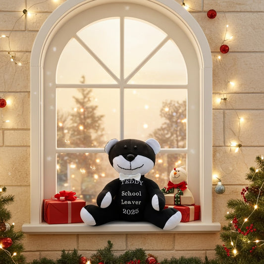 Black and white teddy bear with 'School Leaver 2025' text on a windowsill decorated for Christmas.