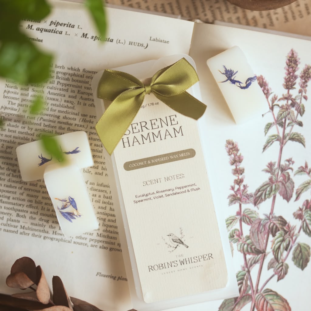 A set of three white scented wax melt bars tied with a green ribbon, displayed on vintage botanical book pages. The centre wax melt is labelled 'SERENE HAMMAM' by 'ROBIN'S WHISPER'.