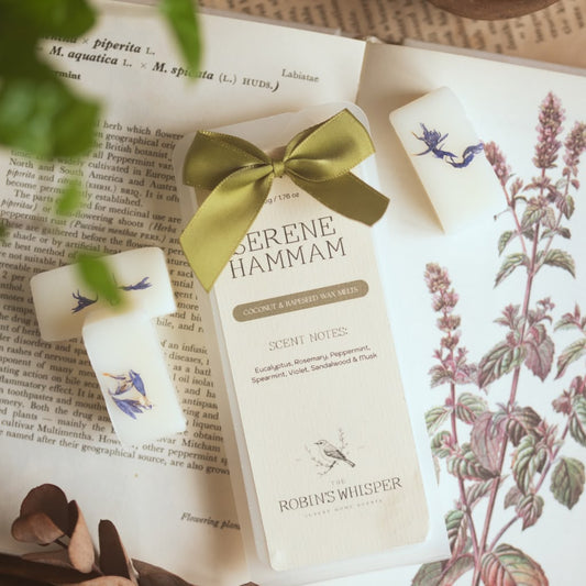 A set of three white scented wax melt bars tied with a green ribbon, displayed on vintage botanical book pages. The centre wax melt is labelled 'SERENE HAMMAM' by 'ROBIN'S WHISPER'.